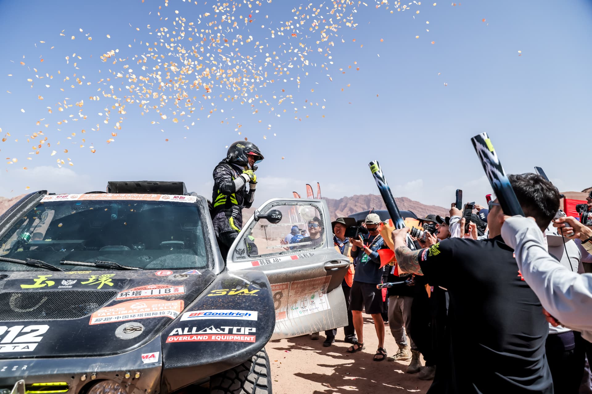 Han Wei standing in the dust-coated HWM rally car as confetti falls at a Taklimakan Rally finish, with photographers alongside