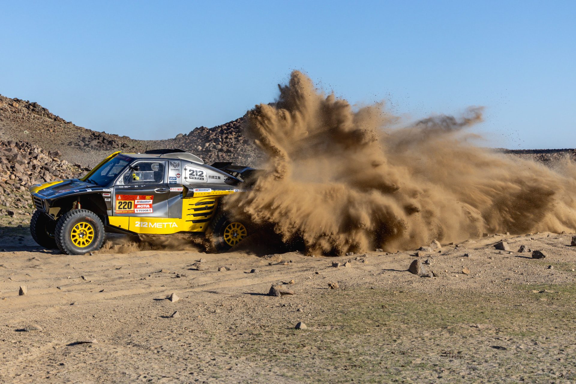 HWM T1+ race car number 220 cornering hard with a tall dust plume against a rocky ridge