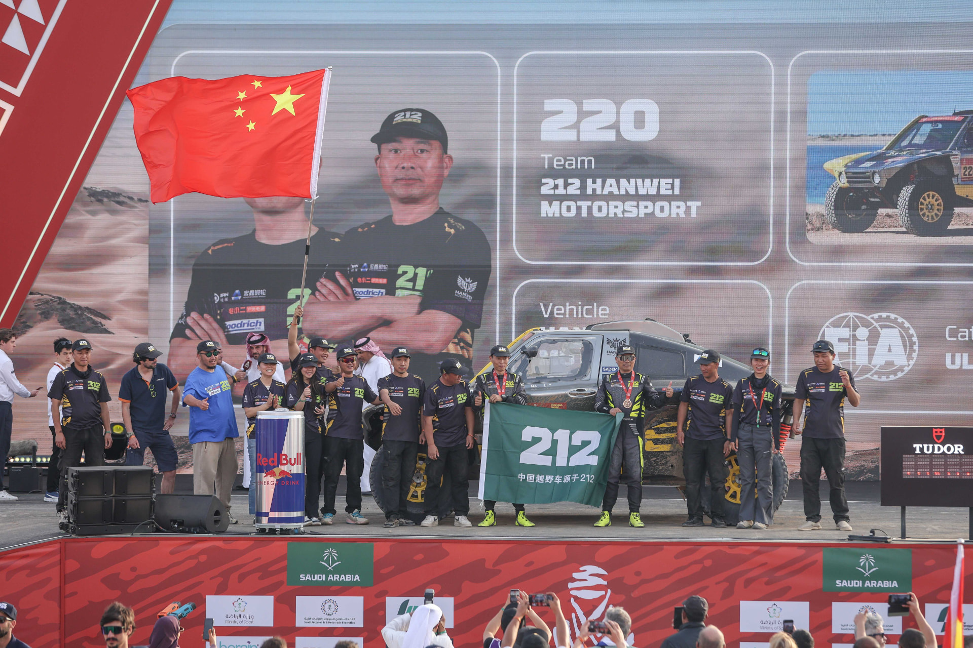 The 212 Hanwei Motorsport team celebrating on the Dakar finish podium in Saudi Arabia, with a Chinese flag and a 212 banner, rally car number 220 behind them